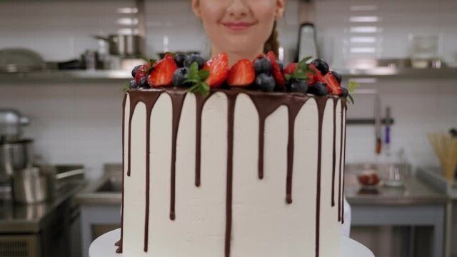 Close-up of a young girl in the uniform of a cook brings a cake with strawberries, chocolate and blueberries to the camera. The pastry chef holds a large creamy cake with strawberries in his hands.
