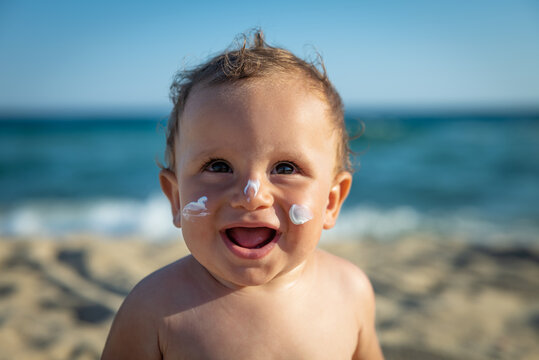 Cute Happy Little Toddler Boy Is Smiling In Camera With Protective Sunscreen Or Sunblock Lotion On His Face Applied By His Mother To Take Care Of Skin On Seaside Beach During Family Holidays Vacation.