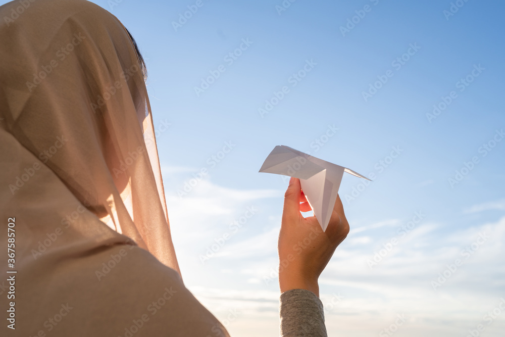 Wall mural silhouette of muslim woman in head scarf launching a paper airplane on blue sunset sky background. t