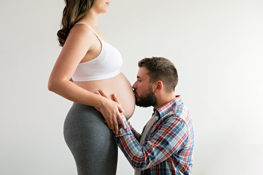 Portrait of bearded man in checkered shirt kissing his wife's pregnant belly over isolated white background. Prenatal period concept. Young family expecting a new child. Close up shot.