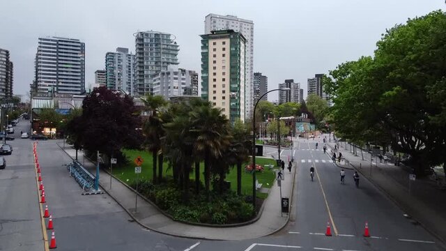 Aerial rise over pandemic restrictive Mortan park tropical trees whereby a Phanotom Drone flys over and criss crosses a bird. Parked cars, cyclists pilan seperation of traffic from Bay Area.