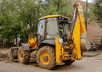 Fototapeta na wymiar Yellow tractor on large wheels with an excavator bucket. Construction machines and equipment for the repair of city sewerage and underground communications.