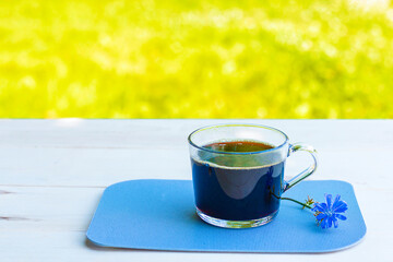 Hot natural chicory caffeine free drink in a transparent cup on a wooden table outdoors. Healthy alternative replacement for coffee, caffeine. Blue chicory flower. Beautiful summer morning at nature
