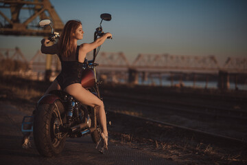 Fototapeta na wymiar Red-haired woman in sexy lingerie in high heels sits on a motorcycle. Attractive red-haired girl sits on a motorcycle at sunset