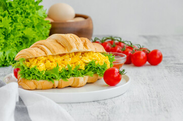 Croissant with scrambled eggs, lettuce and cherry tomatoes on a white plate with ingredients to cook on a wooden background. Horizontal, copy space.