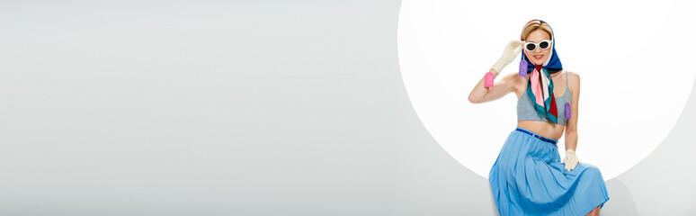 Panoramic shot of smiling stylish woman with price tags on clothes sitting in round hole on white...