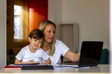 Young Caucasian single mother working on laptop while her son is using digital tablet.Mother and her son spending time together at home.Mother using laptop and her son using tablet.Technology concept.