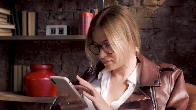 A Young Business Woman Blonde With Glasses Looking Down Using A Smartphone Sitting In A Chair In A Large Bright Office