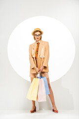 Beautiful woman in straw hat and beige jacket holding shopping bags beside circle on white background