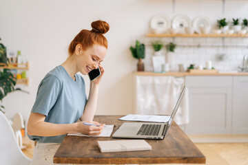 Happy smiling young redhead woman is talking with client by mobile phone, making notes in paper documents, working on laptop. Concept of remote job on Internet.