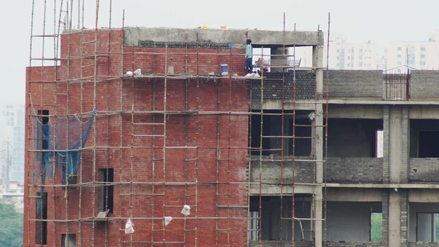 Panning shot from a man working on brick wall bricklaying covering it with cement, standing on a scaffold frame with skyscrapers in the background