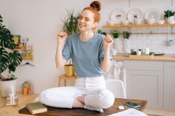 Happy cheerful redhead young woman listening music in wireless earphones while sitting at the desk. Smiling female having fun while listening music with closed eyes at home office.