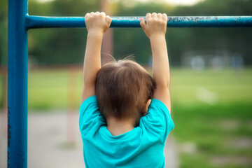 Toddler boy learning to do pull ups: preschooler hanging on worn-out steel bar at sports ground...