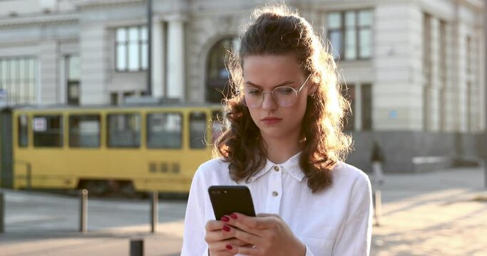 Serious woman in eyeglasses and casual outfit walking on street and using modern smartphone. Concept of everyday routine, busyness and technology.