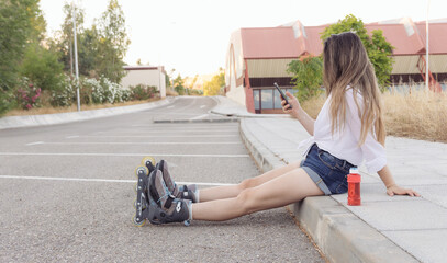 Blonde girl in a white blouse, jeans and skates looking at her mobile phone and sitting in the city at sunset. Roller-skating concept