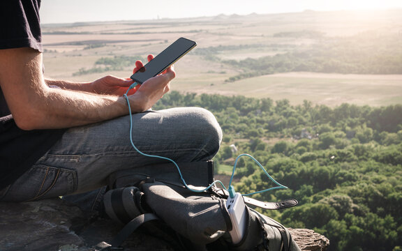 Man on a hike uses smartphone while charging from the power bank on the rock at dawn. Healthy lifestyle and communication.
