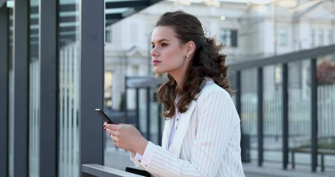 Confident young lady in trendy white suit using modern smartphone for work while standing in business part of big city. Beautiful brunette solving working issues online.