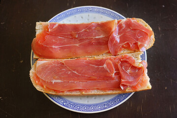 andalusian breakfast with bread, ham and tomato on a dish - picture of a toast of a mediterranean diet from above with a wooden table in the background
