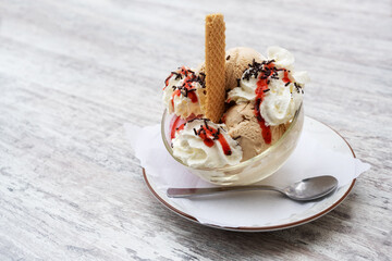 Sundae with ice cream scoops, whipped cream, red fruit sauce and chocolate sprinkles in a glass bowl on a gray table, copy space, selected focus