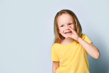 Cute little girl in bright yellow t-shirt holding hand close to mouth sharing rumor or secret. Waist up shot isolated on light blue, copy space