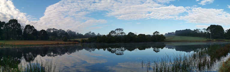 Beautiful morning view of a still pond in a park with stunning reflections of blue puffy sky and...