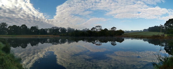 Beautiful morning panoramic view of a still pond in a park with stunning reflections of blue puffy sky and tall trees, Fagan park, Galston, Sydney, New South Wales, Australia