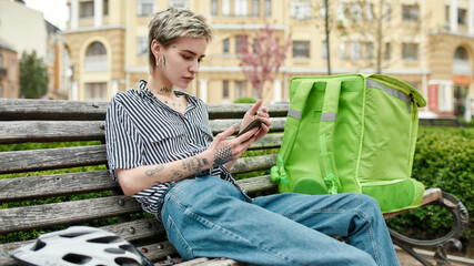 Obraz premium Young delivery woman with thermo bag or backpack checking order using smartphone, sitting on the bench outdoors. Courier, delivery service concept