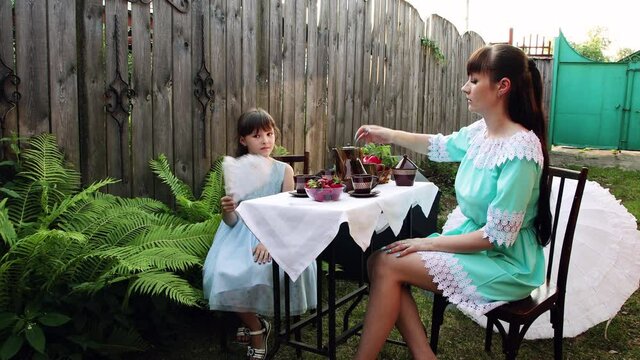 A little girl sits at a table with her mother in the yard. She fanns herself as mom pours tea into a cup. Young ladies in beautiful dresses. Summer day.