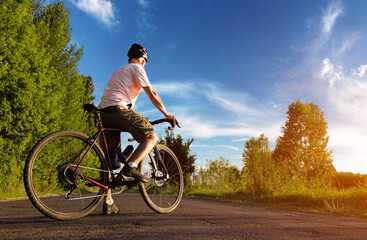 Fototapeta premium Cyclist with bicycle stands on the road at sunset. Man with road bike on the road against the blue sky.