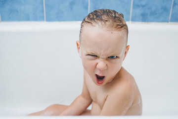 Emotional baby grimaces while washing in the bathroom with foam. about makes a disgruntled wicked...