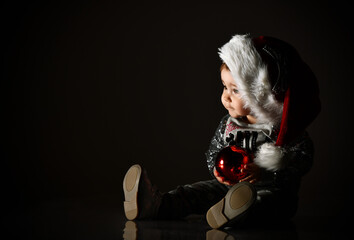 Little female in white hat, gray boots and sparkling suit. Posing sitting on floor in twilight, against black background. Close up