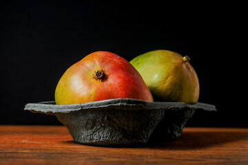 Two fresh whole mango fruit in a black plastic container on a wooden surface, Black background, Fresh fruit retail concept.