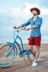 Cool boy with a straw hat on his head is walking on the beach with his blue bike