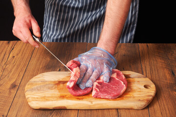 Proffesional butcher in black and white stripes apron, trimming fresh rib eye steak, wooden table and cutting board, Black background, selective focus, Meat industry.