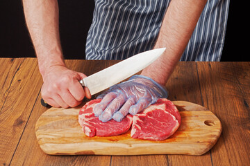 Butcher in black and white apron holding knife in his right hand, his left hand is on a freshly cut rib eye steak, Wooden board and table, black background. Meat industry.