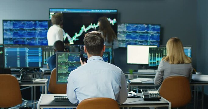 Rear on Caucasian man broker sitting at computer monitor and answering phone call in trading office. Back view on man talking on telephone with client and working with stock market rates and charts.