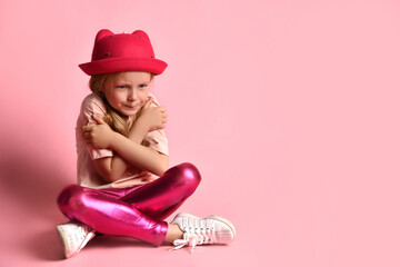 Unhappy little girl in summer clothing and panama hat sitting cross-legged and hugging herself feeling cold. Studio shot isolated on pink, copy space