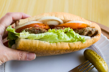 Hand holding of delicious half bread sandwich with beef meatballs,sliced tomato,lettuce and onion rings on wooden plate.Turkish cuisine.