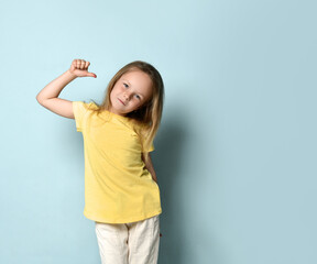 Confident little preschool girl posing in yellow t-shirt pointing with thumbs at herself. Waist up shot isolated on light blue