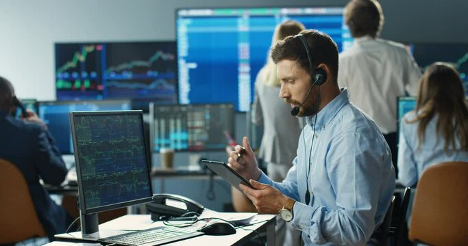 Caucasian man broker in headset sitting in front of computer monitor with charts, graphics and rates and talking with client. Noting in tablet device. Male trader speaking, buying, selling stocks.