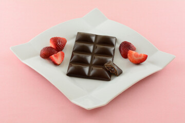 Dark chocolate candy bar with strawberry filling and fresh raw strawberries on white dessert plate on pink background