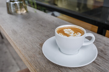 Selected focus and close up view of a white ceramic cup of cappuccino on top with beautiful delicate latte art with leaf pattern, on wooden outdoor rough counter of coffee shop on the sidewalk.