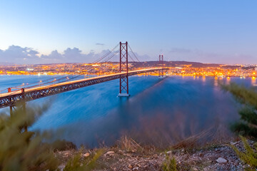 Portugal Travel Destinations. Crossing The Tagus River. Amazing Image of Lisbon Cityscape Along with 25th April Bridge (Ponte 25 de Abril). Taken from Almada District. Picture Taken at Twilight.