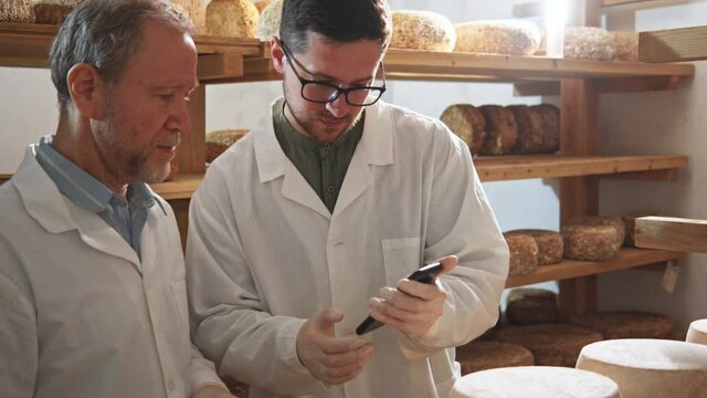 Two farm workers men in white uniform using smartphone at manufacturing. Dairy products online store. Milky farm interior on sunny background. 4K. Technology innovation system. Organic production.