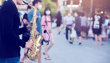 A street musician plays the saxophone and wearing face shield with blurry many people wearing mask...