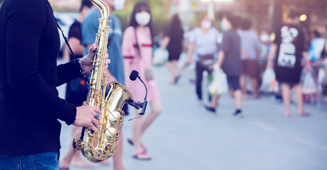 A street musician plays the saxophone and wearing face shield with blurry many people wearing mask...