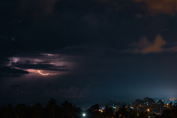 Thunderstorm, lightning and orange clouds in the dramatic night sky over the sea, trees and houses on a tropical island