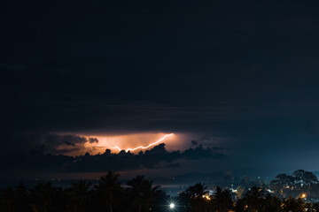 Thunderstorm, lightning and orange clouds in the dramatic night sky over the sea, trees and houses on a tropical island