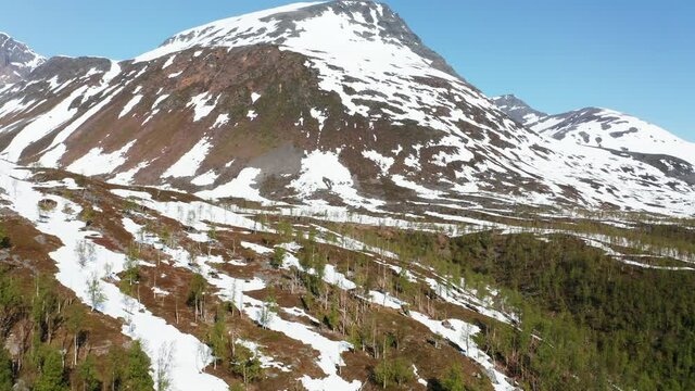Aerial, tilt up, drone shot over trees, towards a snowy mountain peak, in Lyngen alps, sunny, summer day, in North Norway