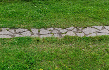 Stone path in the middle of a meadow with green grass. Horizontal track layout. Material - sharp lamellar rock stones.